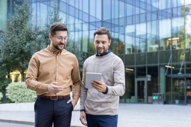Two professional businessmen discussing a new project, collaborating and sharing ideas while looking at a digital tablet screen outside a modern office building