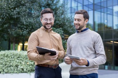 Two smiling male colleagues discussing business and sharing ideas while reviewing information on a digital tablet and clipboard outside a modern building