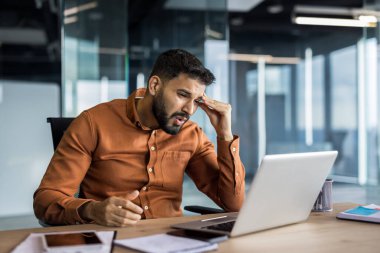 Genç işadamı, modern bir ofiste, masasında oturuyor, bunalmış ve bitkin hissediyor, dizüstü bilgisayarı üzerinde çalışırken acı dolu bir ifadeyle alnını ovuyor.