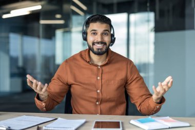 Elleriyle işaret eden kulaklık takan, gülümseyen sakallı bir adam, mükemmel müşteri hizmetleri sunan, çevrimiçi destek veren, ya da modern bir iş ortamında ofis masasından bir webinar yöneten.