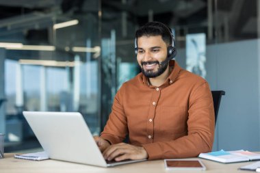 Masada dizüstü bilgisayarda çalışan, mikrofonlu bir kulaklık takan, modern bir çağrı merkezinde veya ofis ortamında çevrimiçi destek sağlayan gülümseyen genç bir adam.