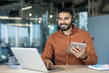 Kulaklık takan ve bilgisayar ve dijital tablet kullanan gülümseyen Hintli adam modern bir ofiste veya uzaktan çalışma ortamında mükemmel müşteri hizmetleri sunuyor.