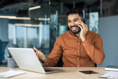 Hintli işadamı cep telefonuyla dizüstü bilgisayar başında gülerek ve el kol hareketi yaparak modern ofis ortamında neşeli bir iş konuşmasının tadını çıkarıyor.