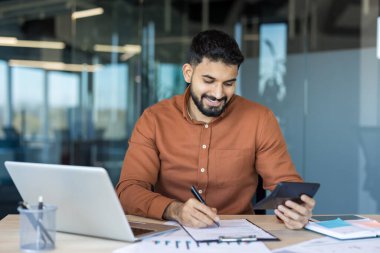 Hintli işadamı masamda gülümserken bilgisayar ve hesap makinesiyle evrakları inceliyor, modern ofis ortamında bütçe, vergi ve finansmanı yönetiyor.