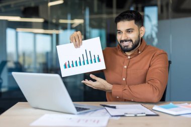 Çevrimiçi bir video konferansı sırasında veri analizi yapan genç işadamı, modern bir ofisten uzaktan çalışırken iş arkadaşlarıyla işbirliği yapıyor.