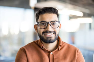 Kameraya güvenle gülümseyen sakallı ve gözlüklü genç Hintli adam modern ofis ortamında başarılı ve ulaşılabilir bir profesyoneli canlandırıyor.