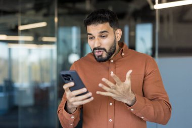 Bir ofiste duran genç adam, elinde akıllı bir telefonla beklenmedik bir kötü haber ya da sürpriz bir mesaj alırken sürpriz, şok ve endişeyi dile getiriyor.