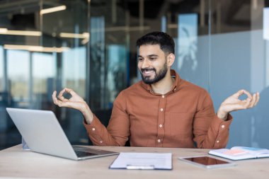 Modern ofis masasında oturan sakallı genç girişimci yoğun bir iş günü sırasında yoga mudra poziyonunda meditasyon yaparak zihinsel huzur ve stres rahatlığı buluyor.