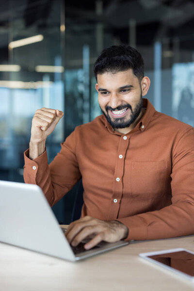 Успешный молодой бизнесмен выражает радость и достижения, работая над ноутбуком в современном офисе, празднуя бизнес-цель или личный успех
