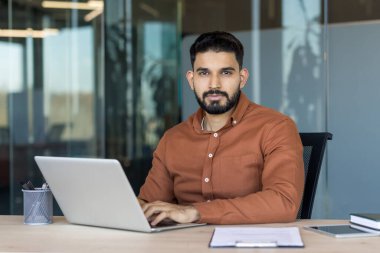 Genç Hintli iş adamı bir masada oturuyor, dizüstü bilgisayarla yazı yazıyor ve kameraya bakıyor, çağdaş ofis ortamında profesyonelliği ve verimliliği temsil ediyor.