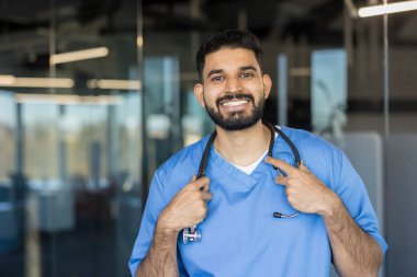 Genç Hintli erkek doktor güvenle gülümsüyor, mavi önlük giyiyor ve steteskop tutuyor, sağlık, ilaç, güven ve profesyonel sağlık hizmetlerini temsil ediyor.