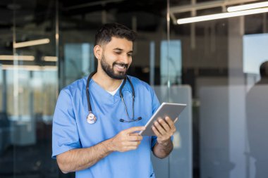 Genç erkek doktor gülümsüyor, önlük giyiyor ve steteskop takıyor, dijital tablet ve teknolojiyle etkileşime geçiyor, modern bir sağlık ofisinde sağlık verilerini yönetiyor.