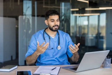 Mavi önlüklü ve steteskoplu Hintli doktor laptoptaki bir hastayla görüntülü görüşme yapıyor, tedavi seçeneklerini açıklıyor ve uzman tıbbi tavsiyeler veriyor.