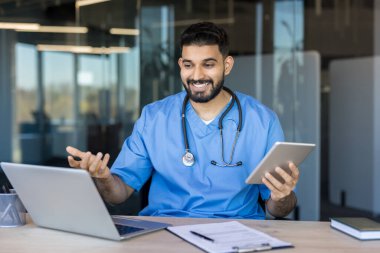 Önlüklü ve steteskoplu bir erkek doktor bilgisayarında bir hastayla görüntülü konuşma yaparken gülümsüyor, elinde tablet ve bir ofisten teletıp hizmetleri sunuyor.