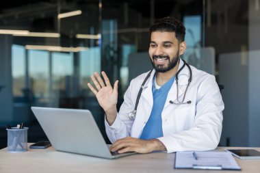 Beyaz önlüklü ve önlüklü Hintli bir erkek doktor çevrimiçi tıbbi konsültasyon yapıyor, bir hastayla telekonferans yapıyor, işyerinden el sallıyor.
