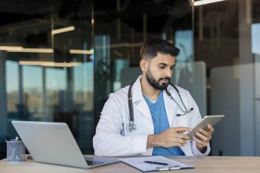 Beyaz önlüklü ve steteskoplu genç erkek doktor bir masada oturmuş, hasta bilgilerini çağdaş ofis ortamında incelerken dijital tablete bakıyor.