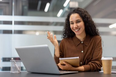 Genç iş kadını bilgisayardaki bir video görüşmesi sırasında gülümsüyor, bir konsepti anlatıyor ve modern bir şirket ofisinde çalışırken notlar alıyor.