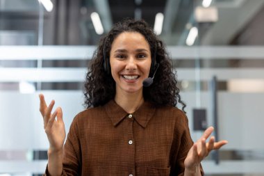 Dost canlısı, kıvırcık saçlı, kulaklık takan, kameraya gülümseyen ve el kol hareketi yapan modern bir ofiste çağrı merkezi operatörü olarak görev yapan bir kadın.