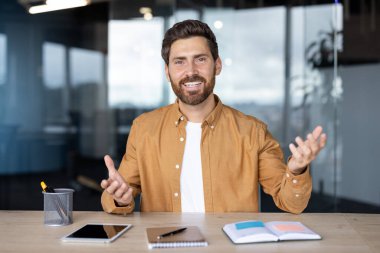 Sakallı, gülümseyen bir adam video görüşmesi yapıyor, modern bir ofiste masa başında otururken iletişim kuruyor ve el kol hareketi yapıyor, online meslektaşlarıyla konuşuyor ya da sunuyor.