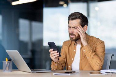 Stresli işadamı cep telefonundan kötü haberler alıyor, modern bir ofiste alnını sıkıca tutuyor, iş ya da mali sorunlarla ilgili endişe, baş ağrısı ve hayal kırıklığı gösteriyor
