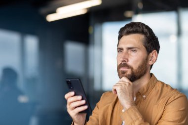 Modern bir ofiste akıllı bir telefon tutan genç düşünceli bir adam, yeni başlayan bir çevrede fikirleri, stratejileri ve gelecek fırsatları düşünürken dikkatle başka tarafa bakıyor.