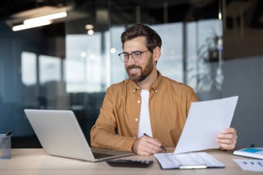Gözlüklü, gülümseyen profesyonel bir adam, modern bir masadaki mali raporları ve evrak işlerini incelerken, kendine güvenen, parlak bir ofis ortamında, dizüstü bilgisayarı üzerinde çalışıyor.