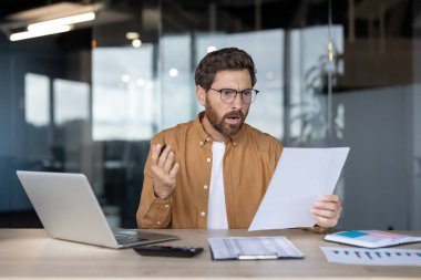 Optik gözlüklü stresli bir adam büro masasında oturuyor, elinde evrak işleriyle, ve bir fatura ya da beklenmedik bir borç gördükten sonra şok olmuş ve kafası karışmış bir ifadeyle tepki veriyor.