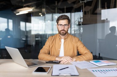 Gözlüklü ve rahat gömlekli kendine güvenen işadamı, elinde laptop, tablet ve belgelerle bir masada oturuyor, çağdaş bir ofis alanında bir portre için poz veriyor.