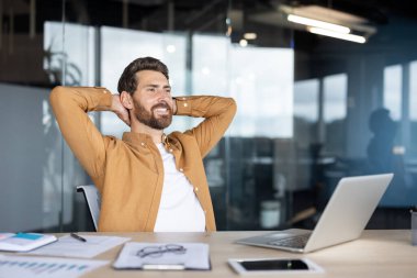 Mutlu iş adamı ofis masasında rahatlatıcı bir moladan zevk alıyor. Ellerini başının arkasına koyup arkana yaslanıyor. Gülümsüyor ve işinden ve başarılarından memnun görünüyor.