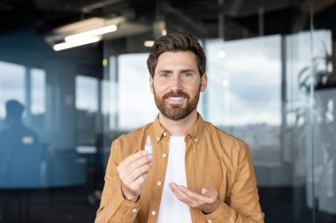 Modern bir ofiste gülümseyen sakallı bir adam, görüş bakımı için göz damlası tutuyor ve sunuyor. Profesyonel yetişkin göz sağlığı, kuru gözler için rahatlama ve günlük göz rutini.