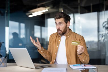 Bilgisayarlı ekranda şok olmuş ve hayal kırıklığına uğramış gibi görünen bir adam, başarısız bir online ödeme veya banka işlemi sonrasında elinde kredi kartı tutuyor, mali bir sorun karşısında şaşırmış hissediyor.