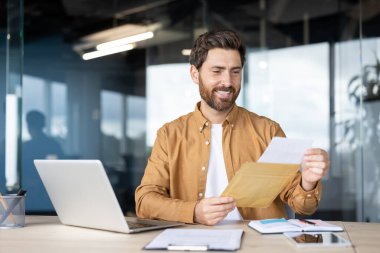 Modern bir ofis masasında oturan mutlu sakallı adam bir zarf açıyor ve bir belge okuyor, başarıyı kutluyor ve olumlu iş iletişiminin tadını çıkarıyor.