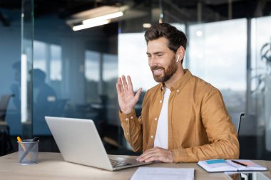 Modern bir ofisten bilgisayarında sanal bir toplantı düzenleyen mutlu sakallı adam gülümseyip el sallıyor, uzaktan çalışma ve dijital iletişimi kucaklıyor.