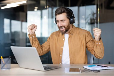 Kulaklıklı mutlu adam bilgisayar ekranına bakıyor ve iş başarısını kutluyor, hedeflere ulaşıyor, ya da online olarak kazanıyor, modern ofiste çalışırken heyecan içinde yumruklarını kaldırıyor.