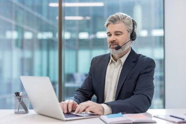 Erkek profesyonel kulaklık takıyor ve dizüstü bilgisayarda yazıyor, müşteri yardımı sağlıyor ve modern ofis ortamından müşterilerle iletişim kuruyor.