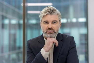 Olgun bir iş adamı, bir ofiste güven içinde poz verirken, doğrudan profesyonelliğin, deneyimin ve idari liderliğin portresini oluştururken çenesini elinin üzerine koyuyor.