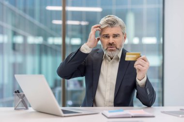 Kafası karışmış bir iş adamı, elinde kredi kartı tutarken kafasını kaşıyor, modern ofis masasında online alışveriş yaparken ödeme sorunları ya da finansal borç sorunları yaşıyor.