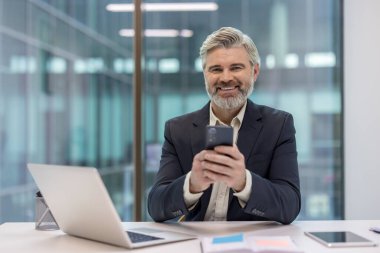 Gri sakallı, gri saçlı, gülümseyen, cep telefonuyla modern bir ofis masasına dizüstü bilgisayarla bakan yetişkin bir adam.