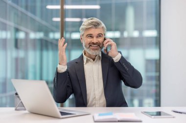 Ofis masasında duran kıdemli işadamı, cep telefonuyla konuşuyor, neşeli bir şekilde gülümsüyor ve eliyle işaret ediyor, kurumsal bir çalışma ortamında etkileşime geçiyor.
