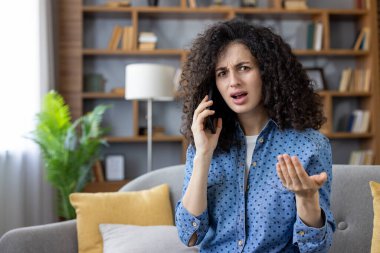 Genç bir kadın, modern oturma odasındaki bir kanepede otururken kızgın ve anlamlı bir telefon konuşması yapıyor, çatışma ve güçlü negatif duygular yaşıyor.