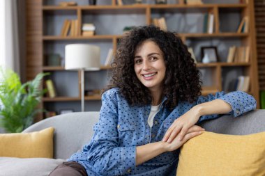 Genç kıvırcık saçlı, mutlu ve kendinden emin bir kadın. Evdeki rahat bir oturma odasında rahat bir kanepede rahatlarken doğrudan kameraya gülümsüyor. Refah ve mahremiyet hissi veriyor.
