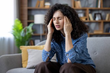 Genç bir kadın kafasında yoğun bir acı ve baskı hissediyor, gözlerini kapatıyor ve ellerini şakaklarına bastırıyor, evdeki bir kanepede oturuyor ve kendini rahatsız hissediyor.