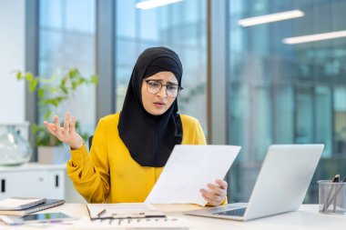 Modern bir ofiste masa başında oturan tesettür ve gözlük takan Müslüman bir iş kadını, kafası karışmış ve hayal kırıklığına uğramış bir halde, kafası karışmış bir ifadeyle evrak işlerine bakarken...