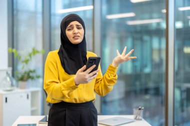 Başörtüsü takan Müslüman iş kadını, bir akıllı telefonu tutarken hayal kırıklığı ve kafa karışıklığıyla tepki veriyor ve kurumsal bir ofiste modern teknolojiyle ilgili kötü haber veya sorun görüyor.