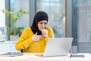 Modern bir ofiste dizüstü bilgisayarı üzerinde çalışırken aniden öksüren Müslüman iş kadını, iş yerindeki hastalık ve sağlık sorunlarını vurguluyor.
