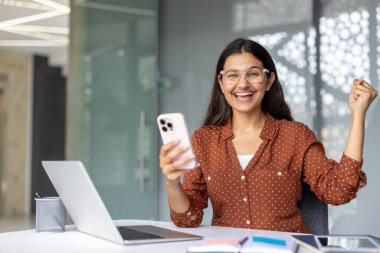 Genç Hintli kadın, akıllı telefonuna bakarken heyecan ve mutluluk ifade ediyor. Modern bir ofiste iyi haber veya başarıyı laptop ve masa başı eşyalar ile kutluyor.