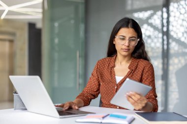 Genç profesyonel bir kadın, masa başında oturmuş, bir yandan bilgisayarla uğraşırken, diğer yandan da modern ofis ortamında dijital tablet içeriğini inceliyor.