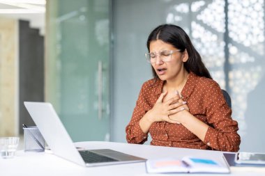 Genç Hintli kadın aniden göğüs ağrısı ya da kalp sorunları yaşıyor. Ofis masasında otururken, iki eliyle göğsünü tutuyor ve rahatsızlık ve ıstırabı ifade ediyor.