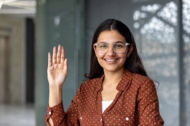 Genç Hintli kadın puantiyeli gömlek ve gözlük takıyor. Gülümsüyor ve el sallıyor. Arkadaşça bir selamlama hareketi yapıyor.