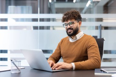 Genç işadamı dizüstü bilgisayarda yazarken gülümsüyor ve çağdaş bir şirket ofisinde güvenle çalışıyor. Mesleki görevlerine ve dijital iş akışına odaklanıyor.